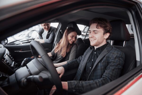 A man, smiling in the driver's seat, and a woman beside him enjoy a "Schedule Test Drives" event while another person stands outside the car, peering in through the open door.