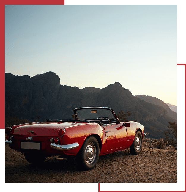 A red vintage convertible car is parked on a dirt road with mountains in the background under a clear sky.